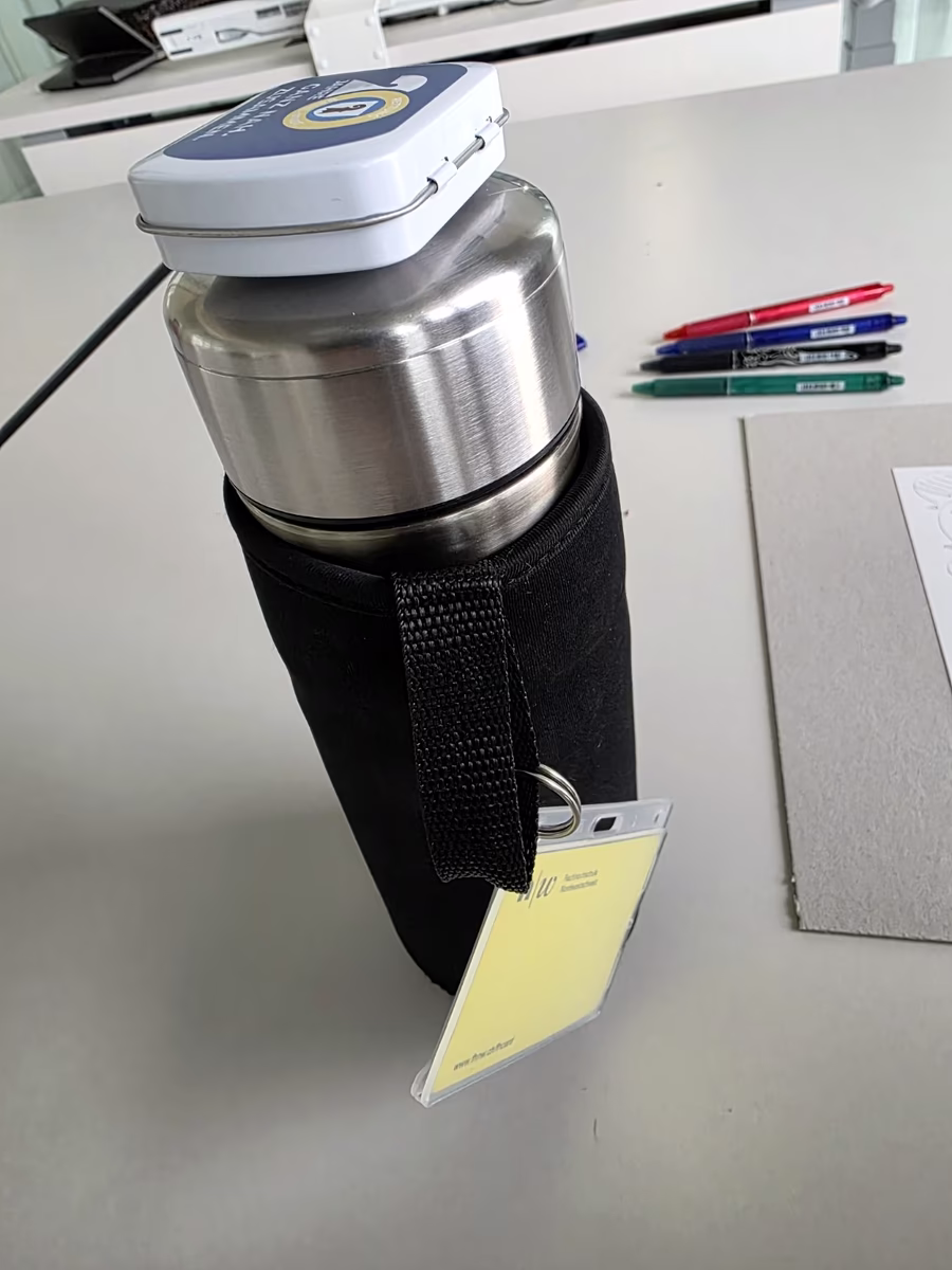 Die Thermosflasche steckt in der Hülle, die Box sitzt oben wie ein Kopf, der Ausweis hängt seitlich wie eine Hand.