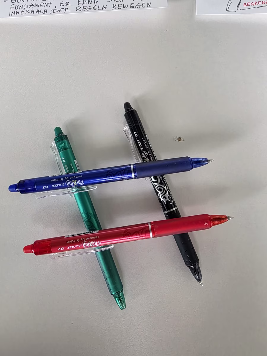 Vier farbige Stifte liegen auf dem Tisch: grün, blau, schwarz und rot.