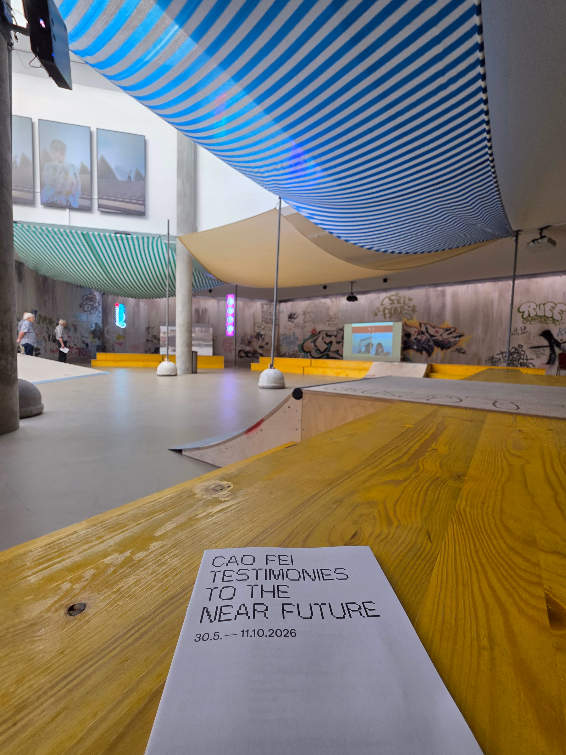 Ein Ausstellungsflyer liegt auf einer gelben Holzfläche. Darüber spannen sich blau und grün gestreifte Stoffbahnen durch den Raum.