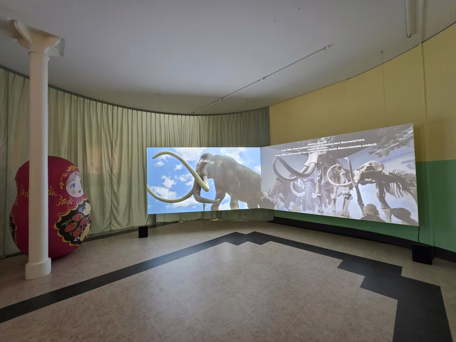 Gebogene Projektionswand mit einem digital rekonstruierten Mammut und fossilen Mammutskeletten, daneben eine übergrosse Matroschka-Figur.
