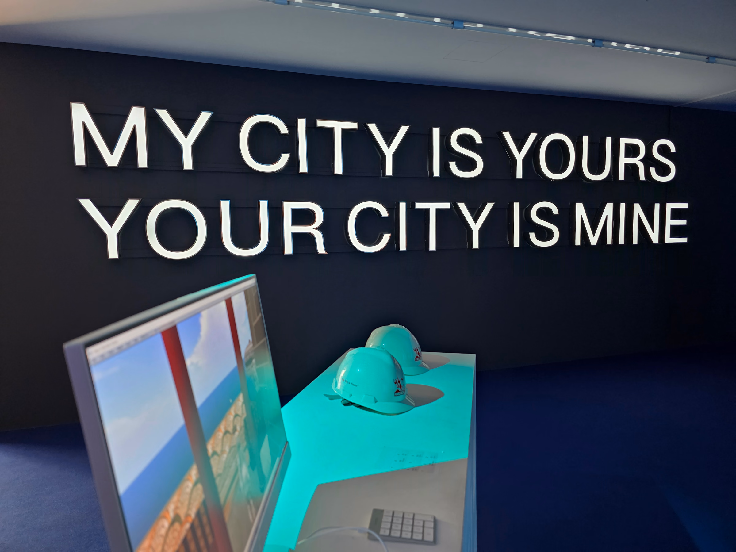 Leuchtschrift MY CITY IS YOURS / YOUR CITY IS MINE an einer dunklen Wand, davor zwei Bauhelme und ein Bildschirm mit einem virtuellen Stadtmodell.