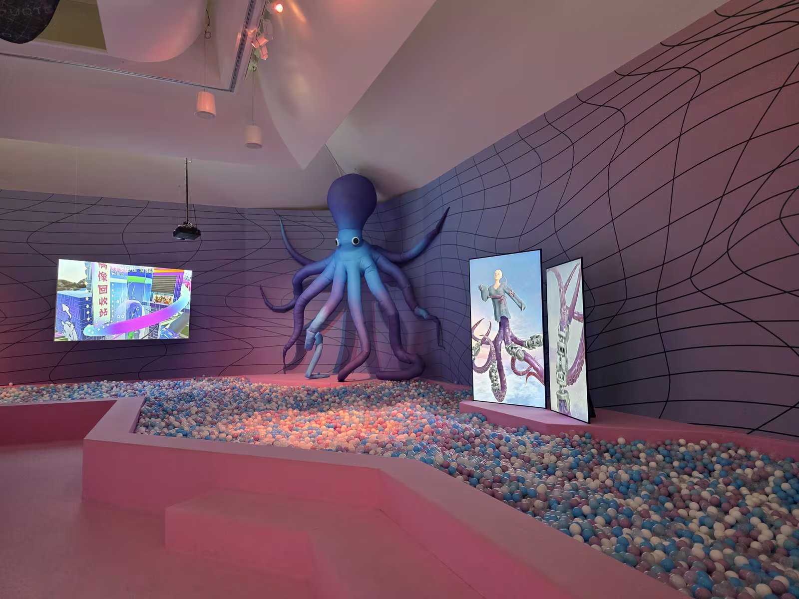 Ein aufgeblasener blauer Oktopus über einem Ballbad aus rosa, blauen und weissen Kugeln, daneben Bildschirme mit einer virtuellen Stadt und einem Avatar.