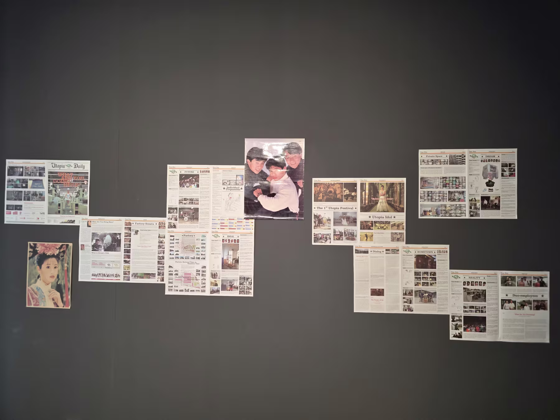 Mehrere unregelmässig an eine dunkle Wand geheftete Zeitungsseiten der fiktiven Publikation Utopia Daily, dazwischen Porträtfotos.
